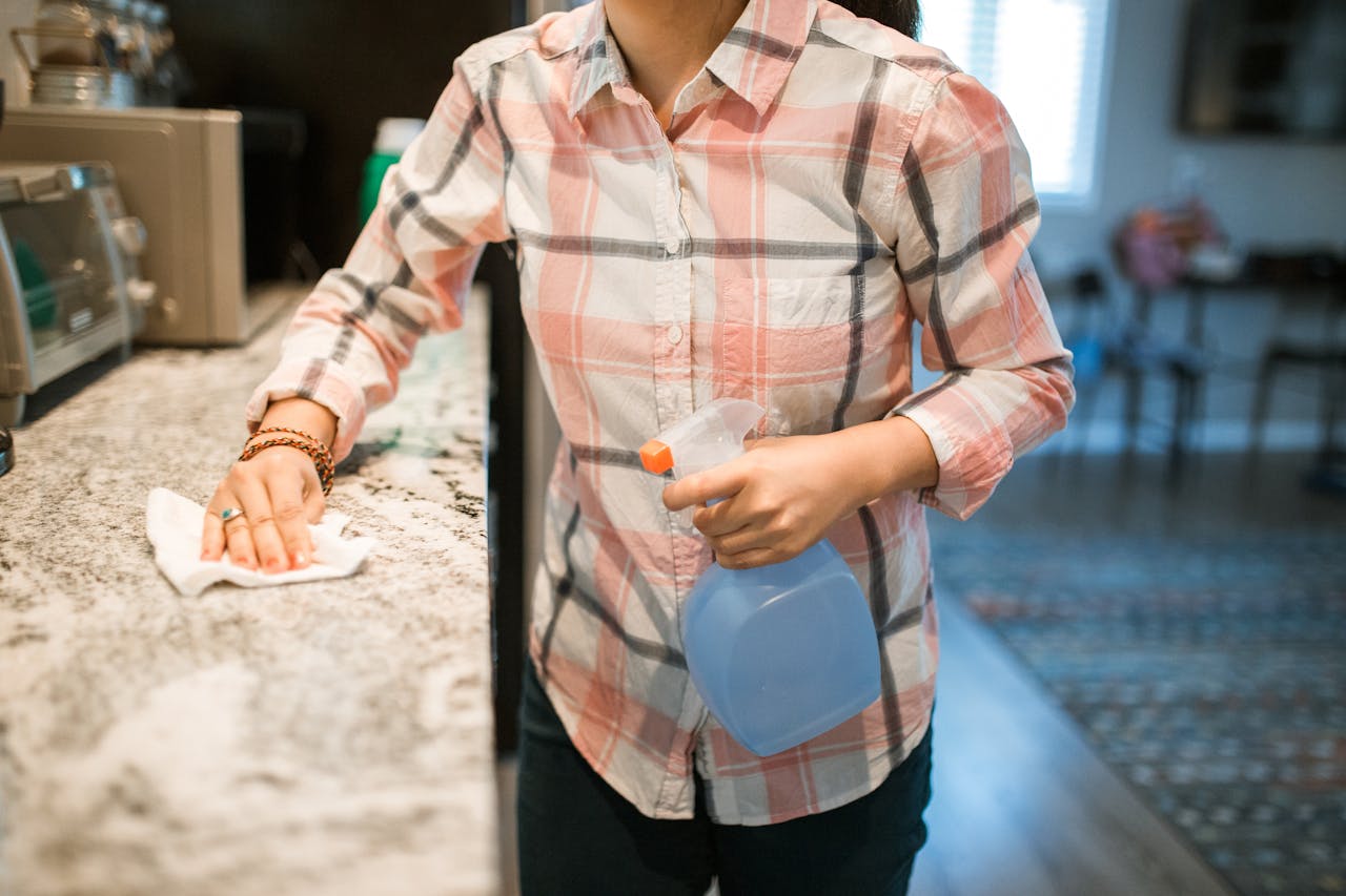 expertise-01 A woman wearing a plaid shirt cleans a kitchen counter using a spray bottle and cloth.